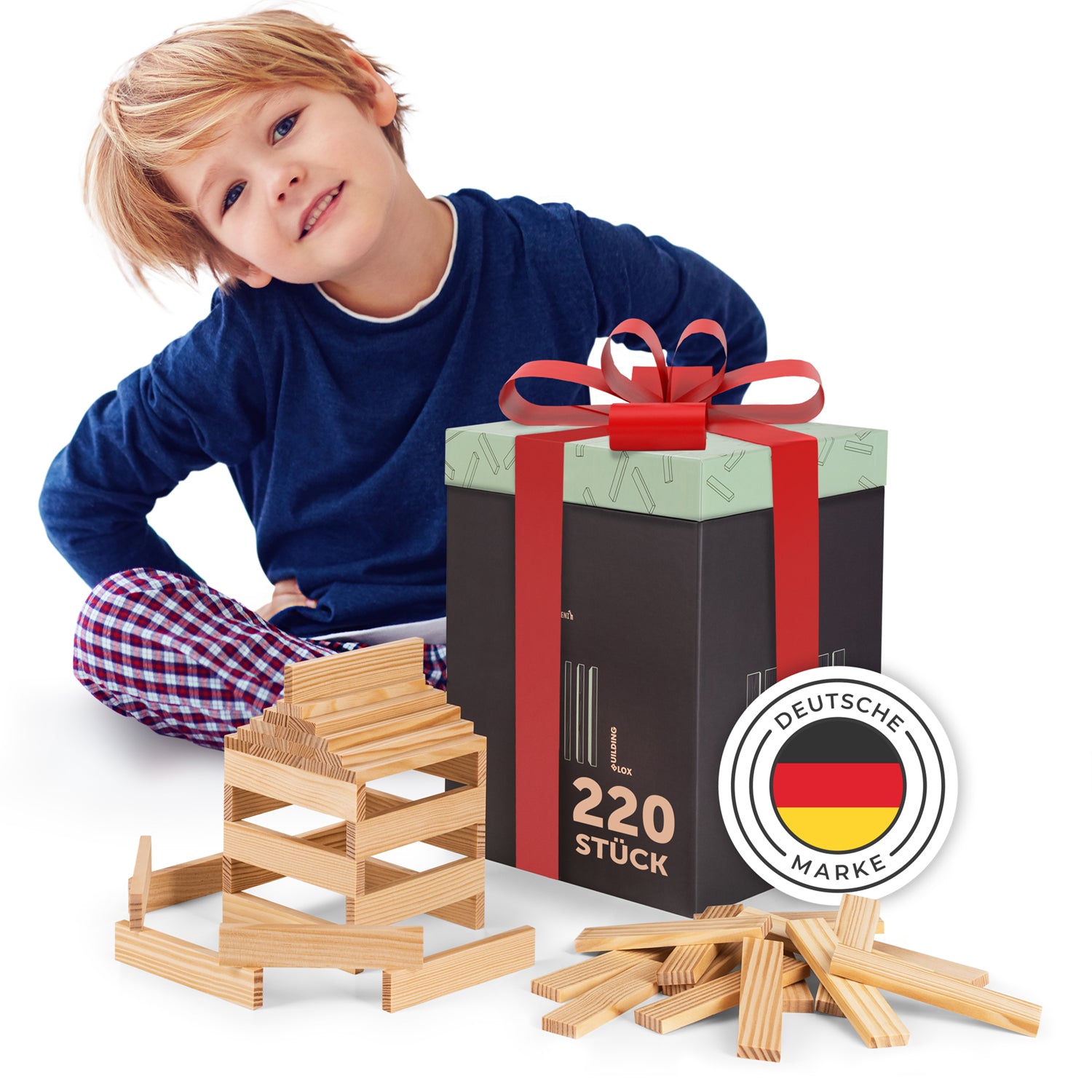 Junge mit blauem Oberteil und karierten Hosen sitzt neben einem Geschenk und einer Konstruktion aus Holzklötzen, Markenzeichen mit deutscher Flagge ist sichtbar.