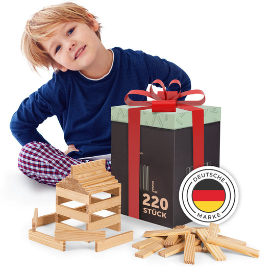 Junge mit blauem Oberteil und karierten Hosen sitzt neben einem Geschenk und einer Konstruktion aus Holzklötzen, Markenzeichen mit deutscher Flagge ist sichtbar.