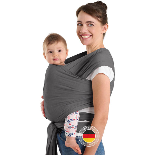 Frau mit Hochsteckfrisur trägt Baby in grauer Tragehilfe vor sich beide schauen zur Kamera.