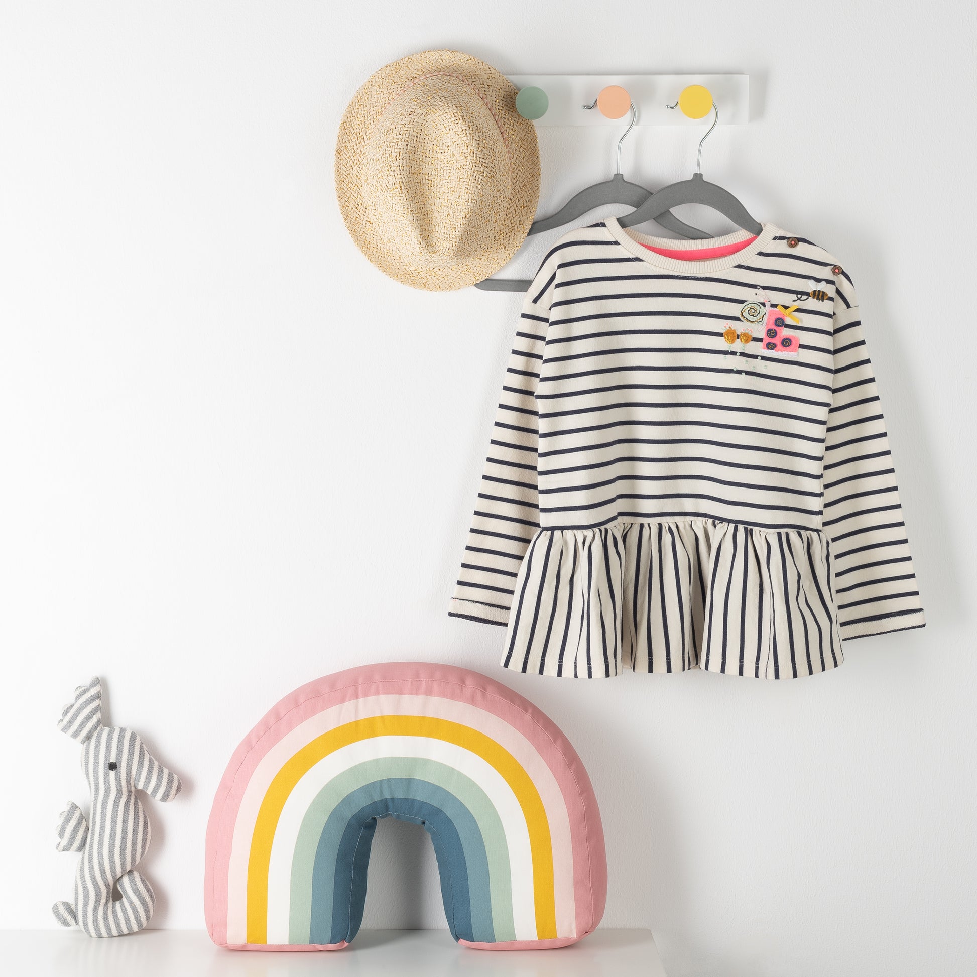 Wandhaken mit Strohhut und gestreiftem Kinderkleid auf grauen Kinderkleiderbügel neben gestreiftem Kissen in Regenbogenfarben