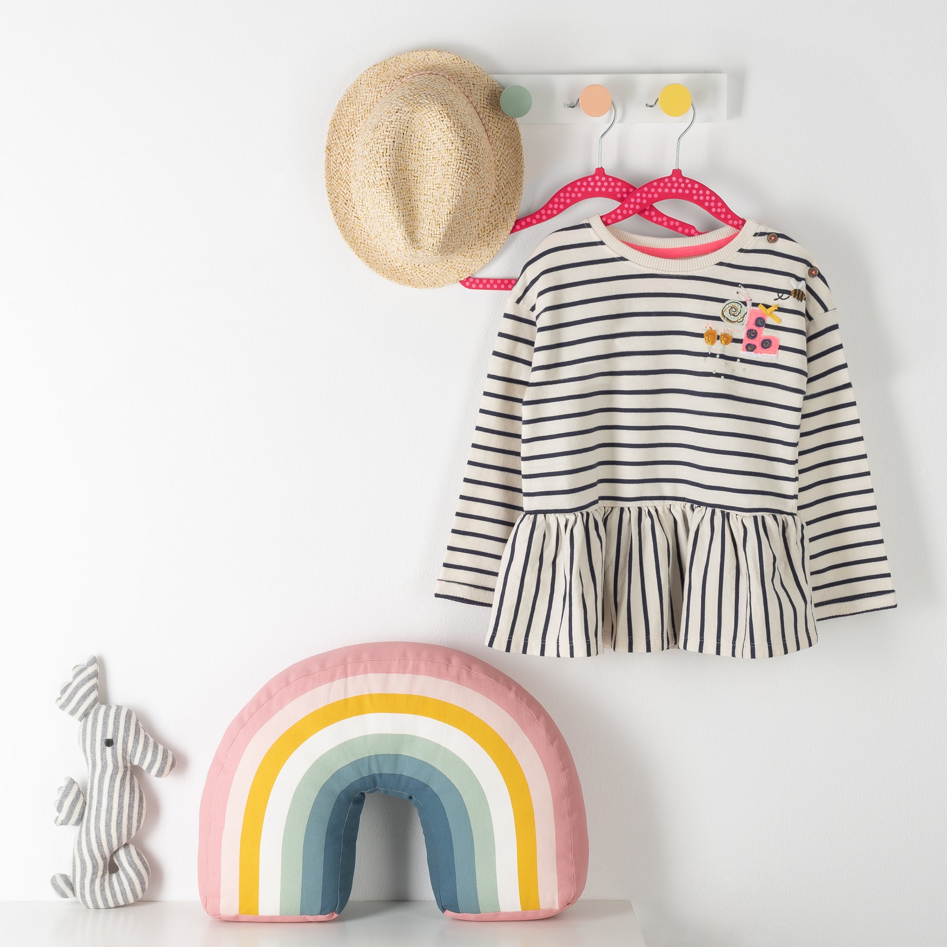 Wandhaken mit Strohhut und gestreiftem Kinderkleid auf pinkem Kinderkleiderbügel neben gestreiftem Kissen in Regenbogenfarben