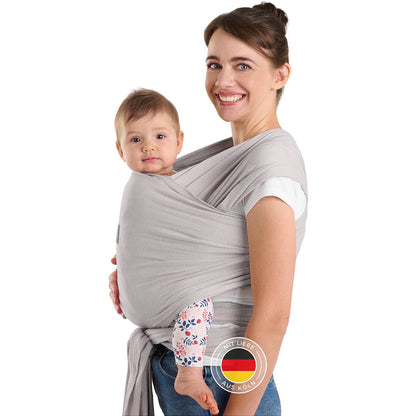 Frau mit Hochsteckfrisur trägt Baby in hellgrauer Tragehilfe vor sich beide schauen zur Kamera.